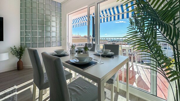 Een eetruimte met een tafel gedekt voor twee personen, een uitzicht op de stad door grote ramen en een plant in pot op het balkon.