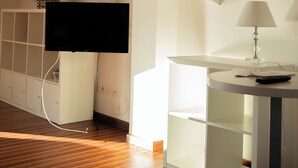 A room with a wooden floor, a flat-screen TV mounted on the wall, a white shelving unit, and a table with a lamp. - Comfortable Living Space in Sorrento (Sant'Agnello)