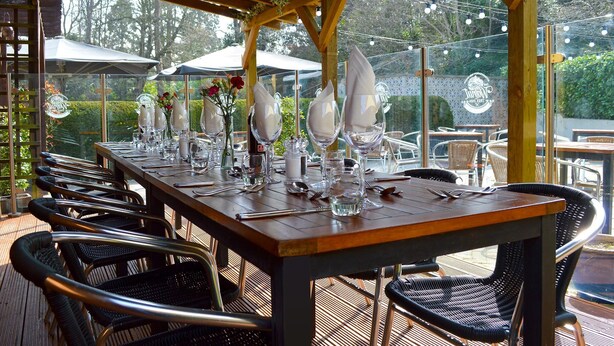 Un área de comedor al aire libre con una larga mesa de madera dispuesta para una comida, rodeada de sillas de mimbre.