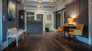 Un pasillo amplio con suelo de madera, un banco blanco, una zona de comedor con mesa y sillas, y un mostrador de recepción con una planta.