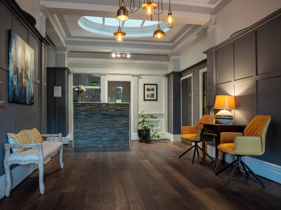 A spacious hallway with a wooden floor, a white bench, a dining area with a table and chairs, and a reception desk with a plant.