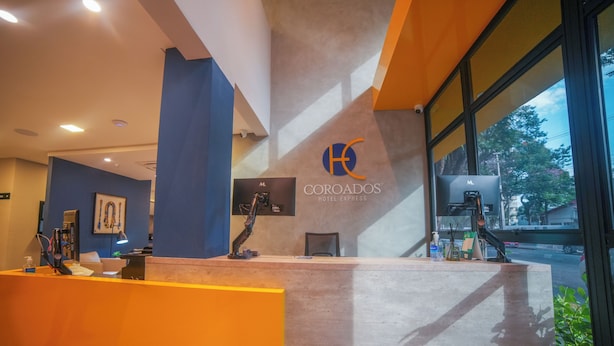 A hotel reception desk with a modern design, featuring a prominent logo and a view of the outside through large windows. - Hotel Coroados Foz Express (Foz do Iguaçu)