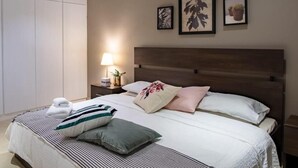 A neatly made bed with a striped blanket, pillows, and a folded towel. There is a bedside table with a lamp and a small plant. The room has a wooden headboard and a large wardrobe. - Nakheel Residence by House living (Sabah Al Salem)