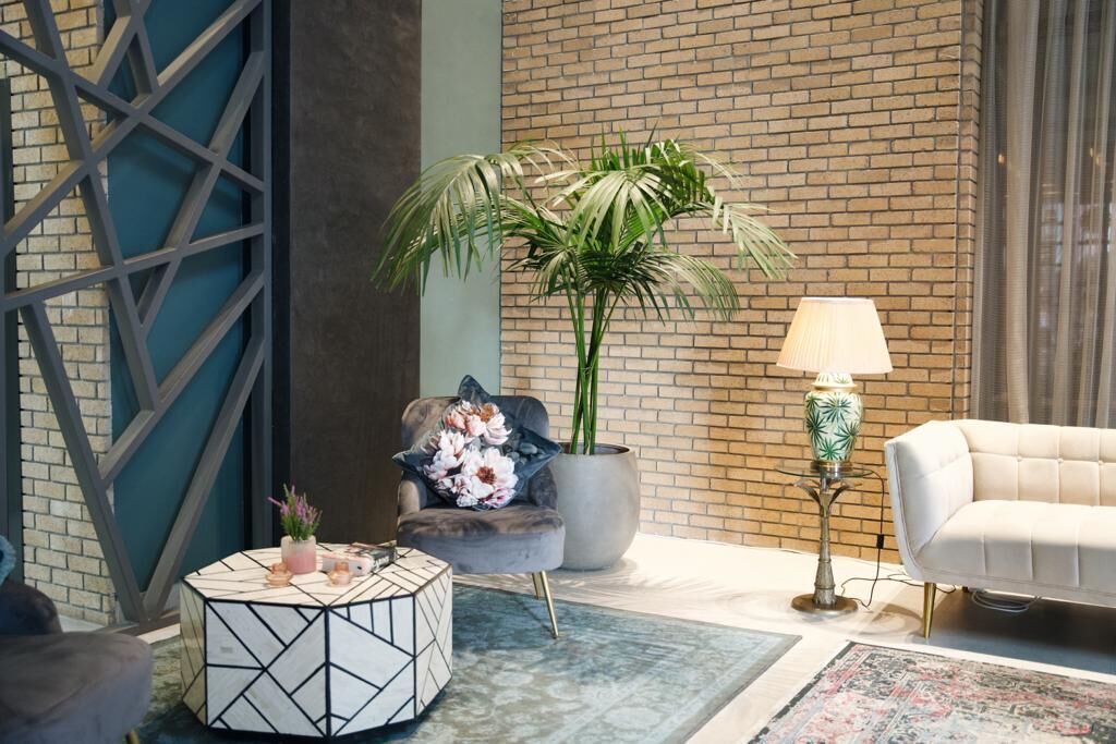 Una sala de estar moderna con pared de ladrillo, una mesa de centro redonda con diseño geométrico, un sillón gris con cojín floral, una planta grande en maceta, un sofá blanco y una lámpara de pie.