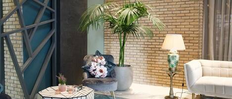 Una sala de estar moderna con pared de ladrillo, una mesa de centro redonda con diseño geométrico, un sillón gris con cojín floral, una planta grande en maceta, un sofá blanco y una lámpara de pie.