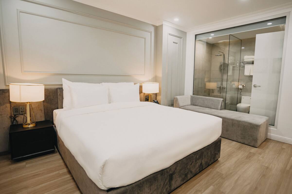 Una habitación de hotel moderna con una cama grande, lámparas de noche, un sofá, una ducha envuelta en cristal y un vestidor.