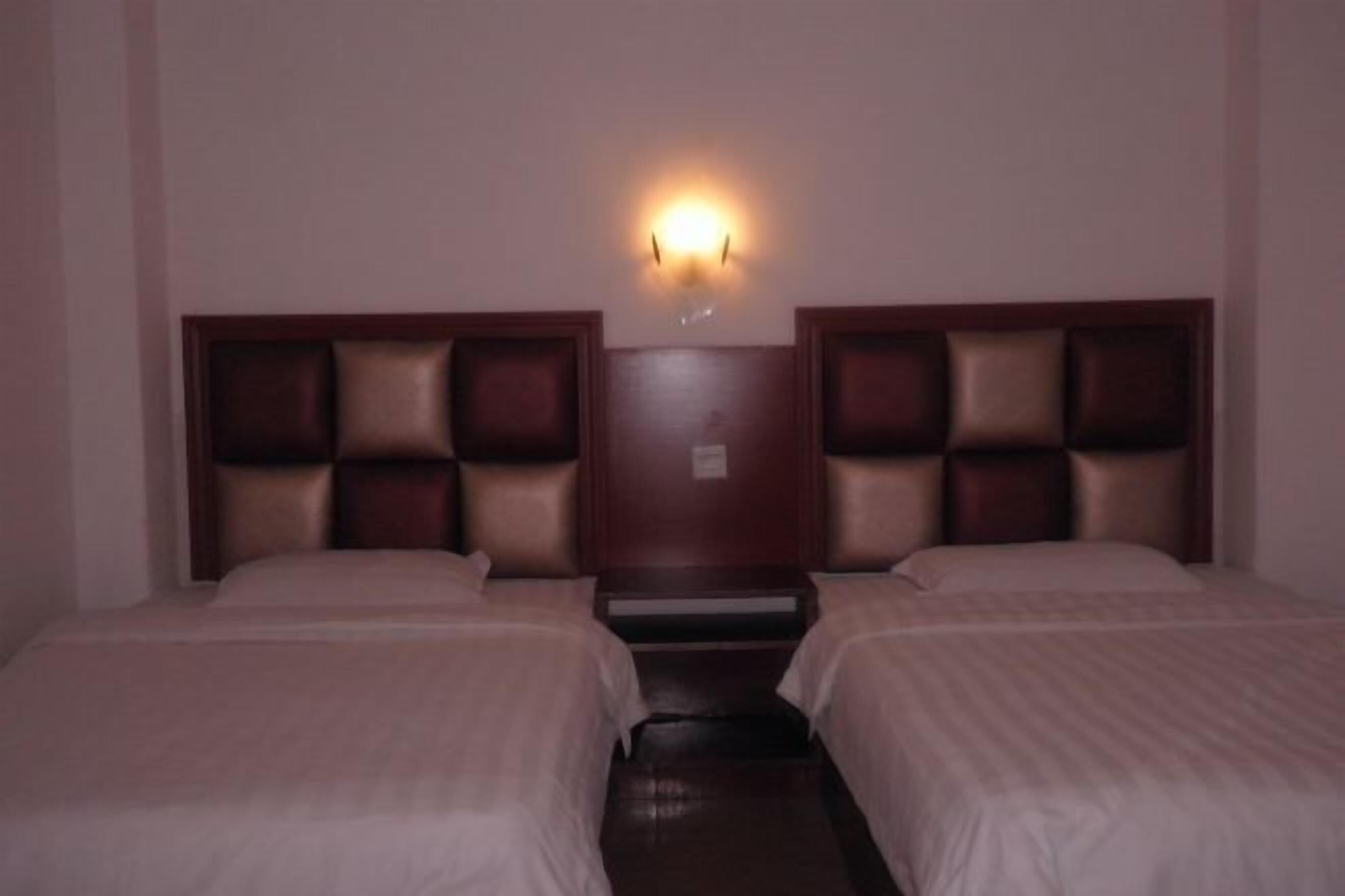 Quarto de hotel com duas camas, uma mesinha de cabeceira central e uma luminária fixada na parede.