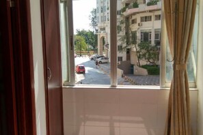 Une vue depuis une fenêtre, à travers des rideaux, donnant sur une rue où sont garées des voitures, avec des bâtiments en arrière-plan.