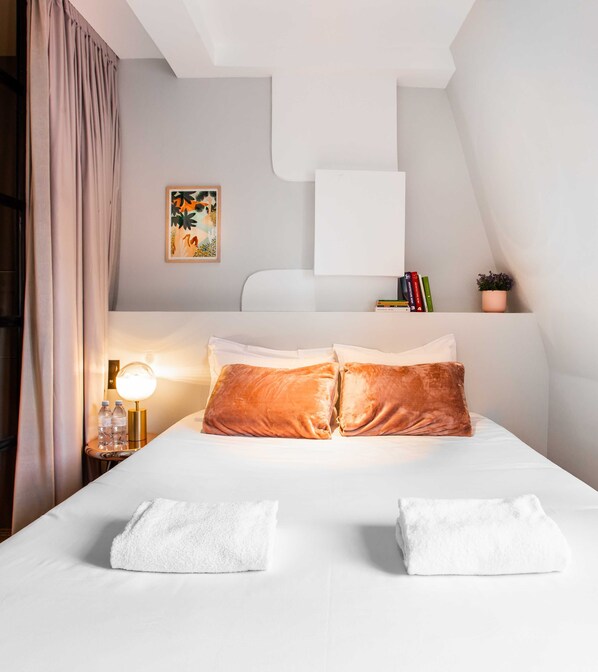 A neatly made bed with two pillows, a bedside lamp, a small table with water bottles, a framed picture, and a potted plant on a white wall. - Résidence Paris 14 CAIRE (Paris)