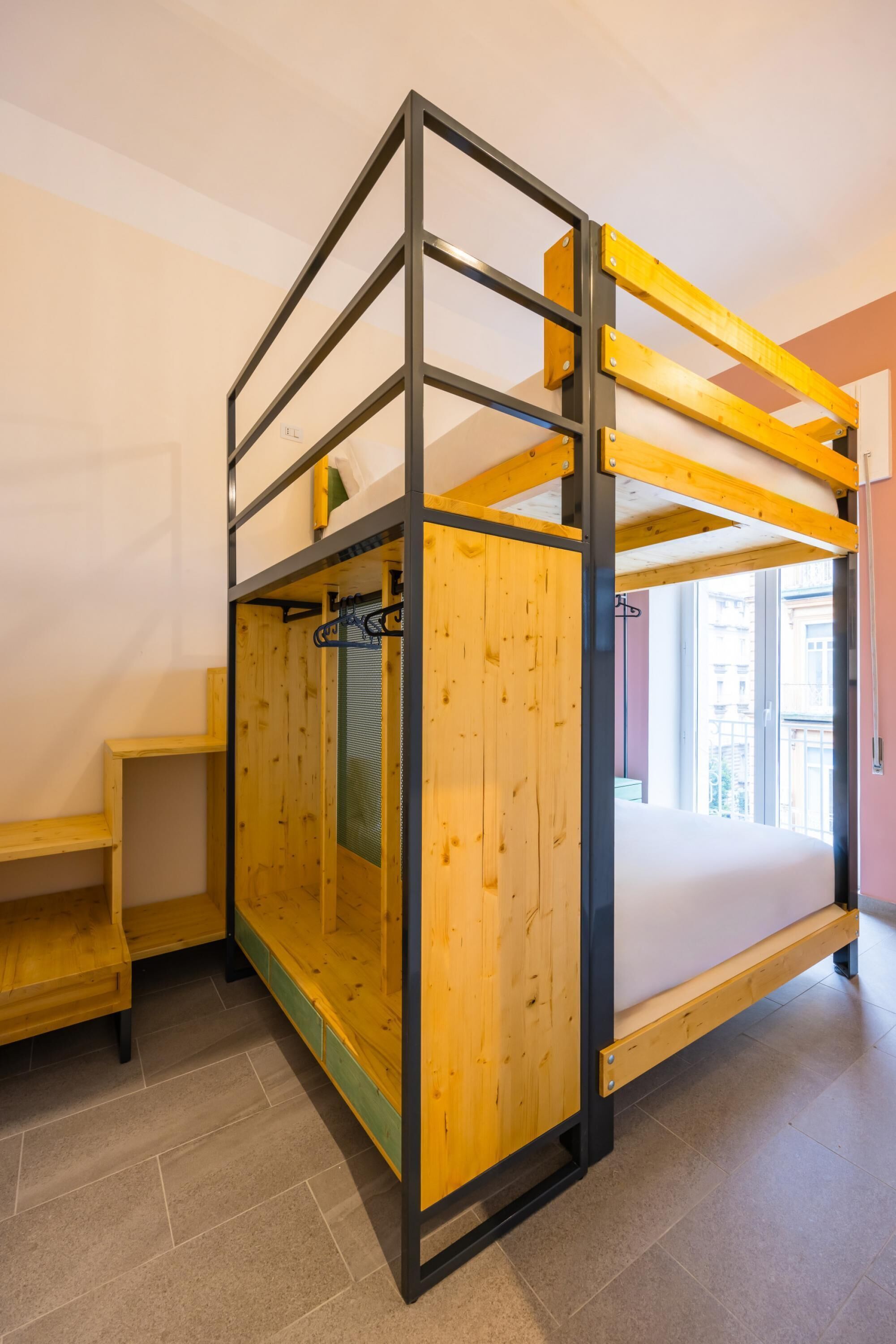 Una habitación con literas, muebles de madera, un ventanal y vistas a edificios.