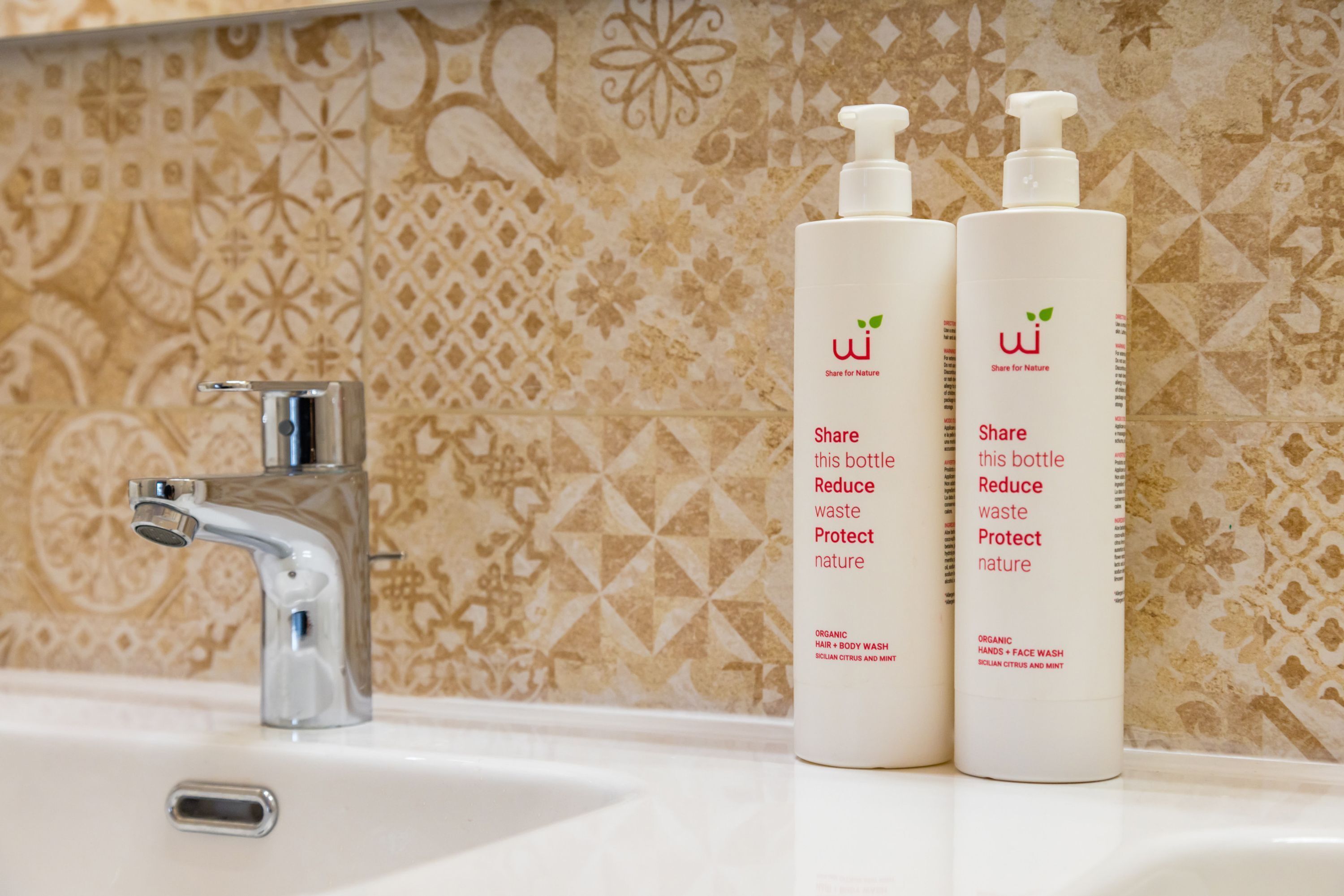 A bathroom with a sink, two bottles of organic hand and body wash, and a decorative tiled wall.