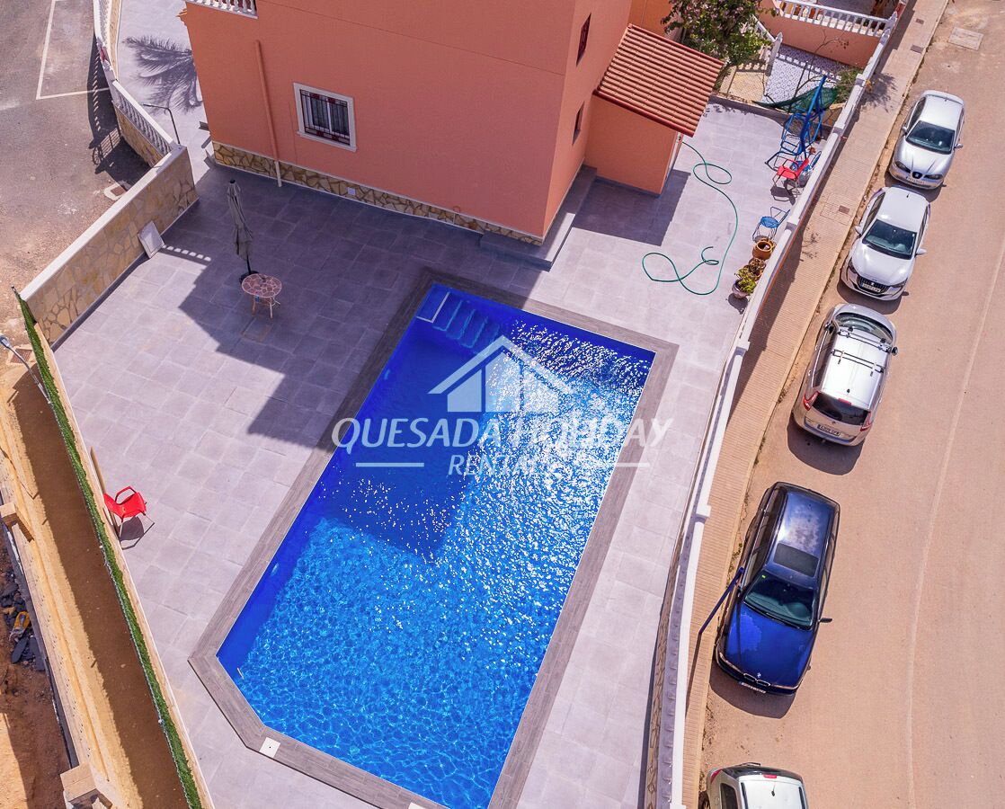 Une piscine avec un élément décoratif aquatique aux eaux bleues, située à côté d’un bâtiment à la façade rouge et d’un espace de stationnement où sont garées plusieurs voitures.