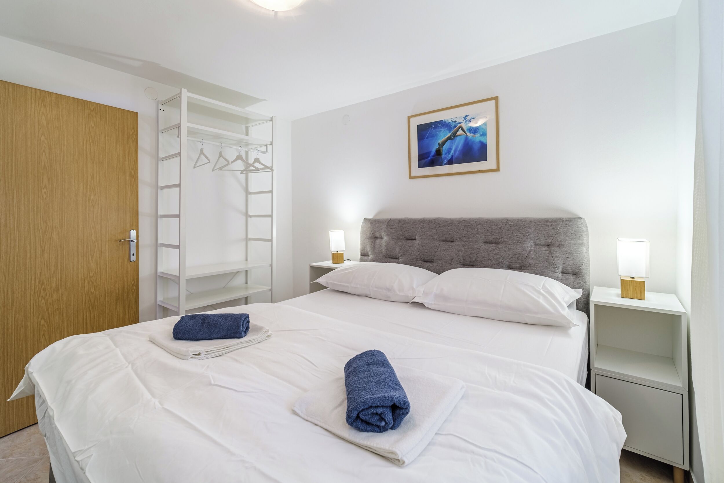 Un lit bien fait, avec du linge de lit blanc et deux serviettes enroulées, une tête de lit grise, une table de chevet blanche avec une lampe, une armoire en bois et un tableau encadré au mur.