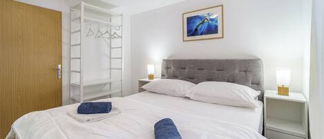 Un lit bien fait, avec du linge de lit blanc et deux serviettes enroulées, une tête de lit grise, une table de chevet blanche avec une lampe, une armoire en bois et un tableau encadré au mur.