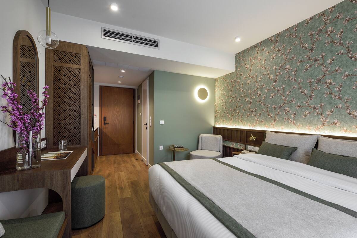 A modern hotel room with a large bed, a desk with a mirror, a round wall light, and a floral wallpaper.