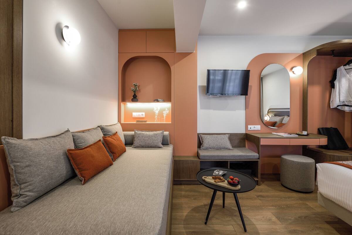 A modern hotel room with a sofa, a small table with snacks, a desk, and a mirror.