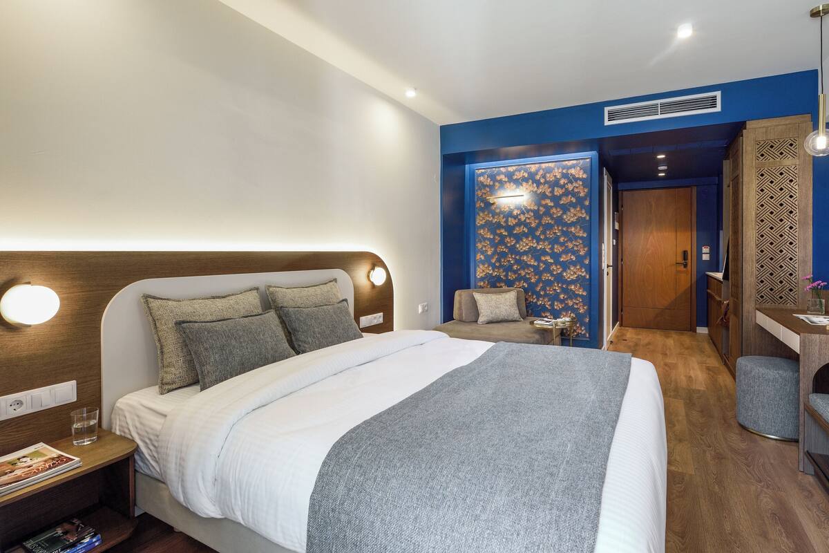 A modern hotel room with a large bed, bedside table, and a desk with a chair.