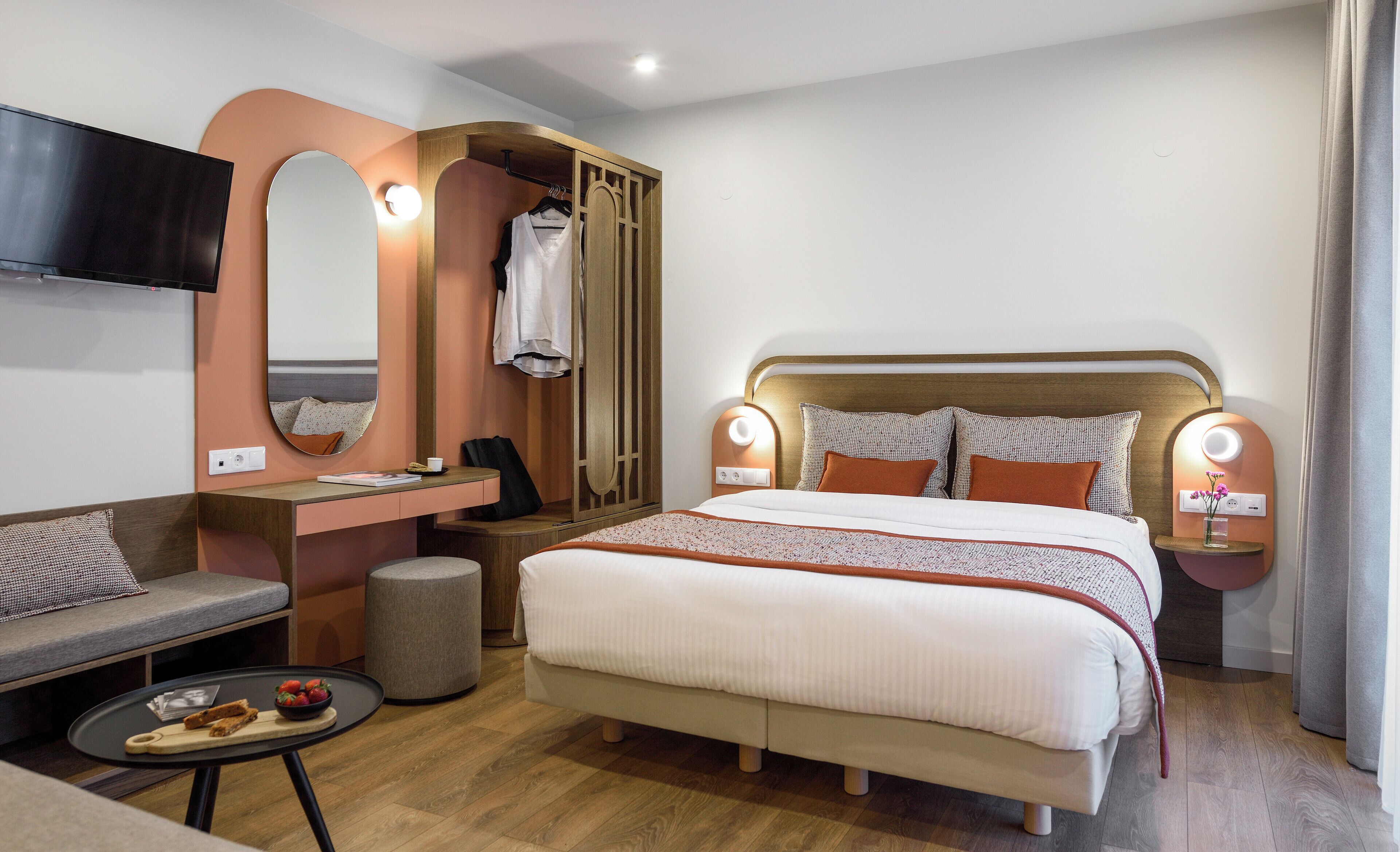 A modern hotel room with a large bed, a seating area with a sofa and a small table, a vanity with a mirror, and a television mounted on the wall.