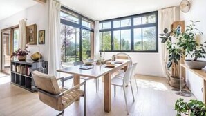 A dining area with a wooden table and chairs, large windows, and indoor plants. - Pomerolle - Saoû - 4 pers (Saou)