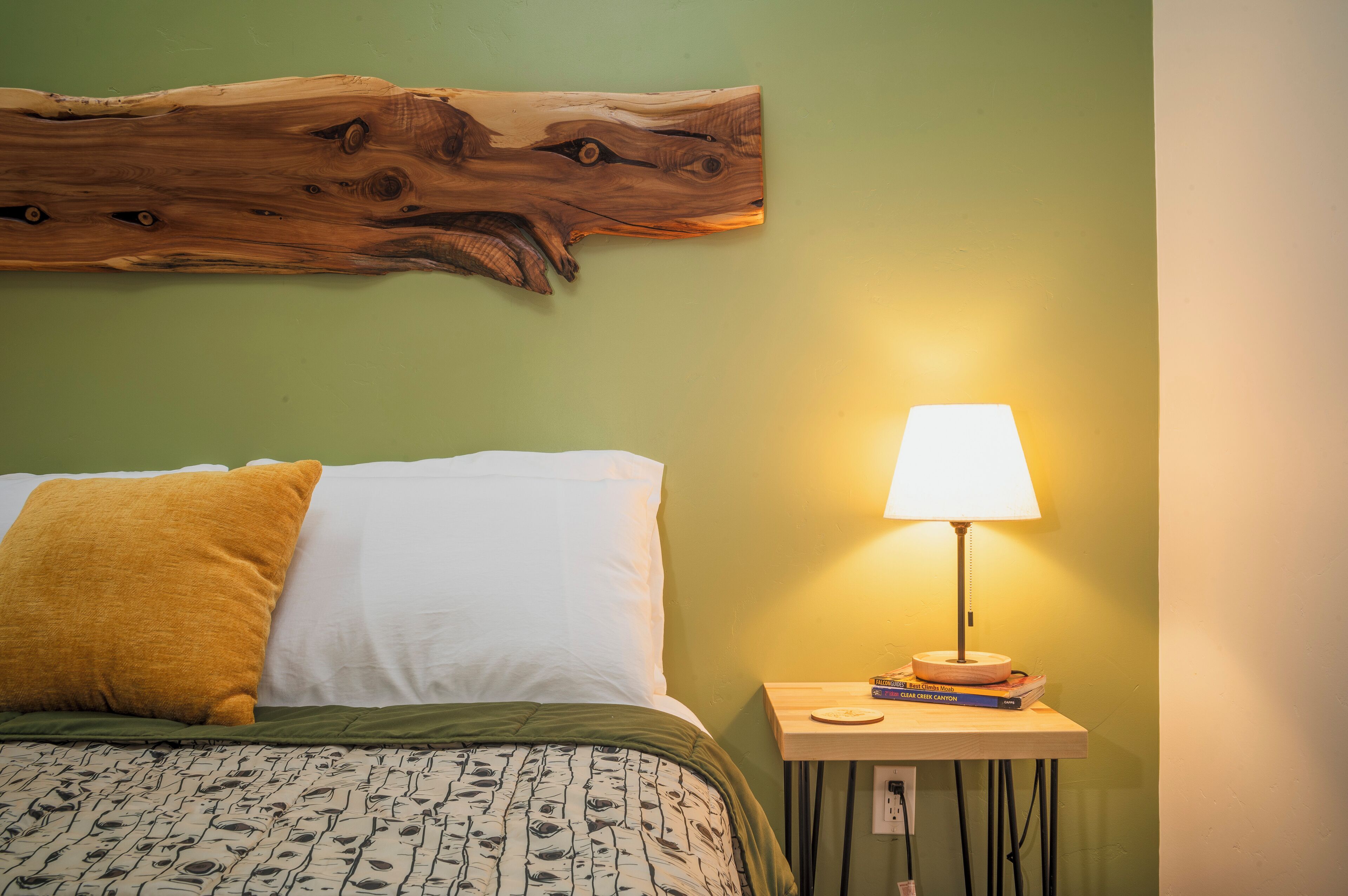 Una cama con una colcha estampada, un cabecero de madera, un almohadón amarillo, un almohadón blanco y una mesita de noche con una lámpara y libros.