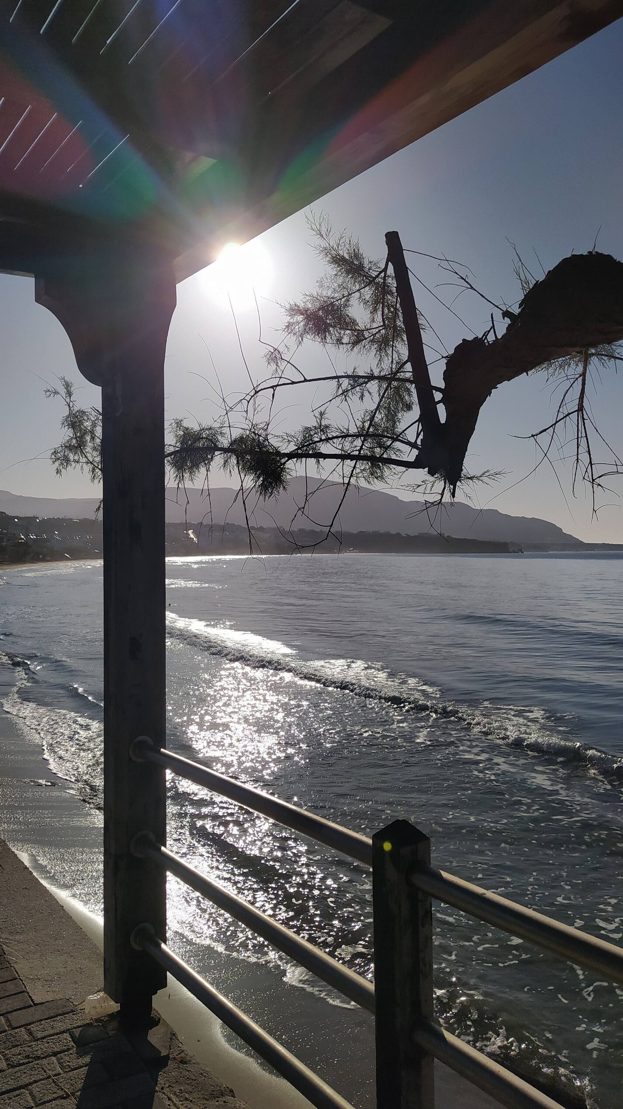 Blick auf die Küste mit einem Metallgeländer, blauem Himmel und Sonnenlicht, das sich auf dem Wasser spiegelt.