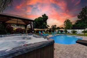 A hot tub and a swimming pool with lounge chairs, surrounded by trees and a sunset sky.