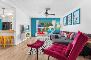 A modern living room with a grey sofa, a red chair, and a kitchen area with a fridge and bar stools.
