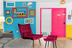 A room with a vibrant blue wall, a red velvet chair, a small ottoman, a bright pink door, and a window with blinds. - Happy Hacienda- Heated Pool, Hot Tub, Golf, Outdoor Games (Bradenton)