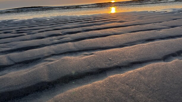 Ein Strand mit Wellen im Sand und einem Sonnenuntergang im Hintergrund.