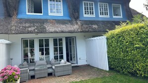 Ein Haus mit einem Reetdach, blauer Fassade und einer Terrasse mit Korbmöbeln.