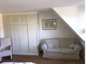 A room with a white wardrobe, a checkered sofa, a white armchair, and a framed picture on the wall.