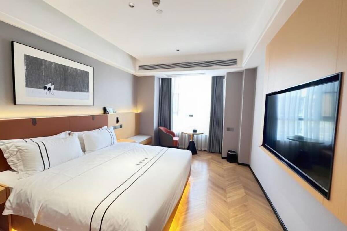 Una habitación de hotel con una cama grande, un televisor de pantalla plana, un cuadro en la pared y una ventana con cortinas.