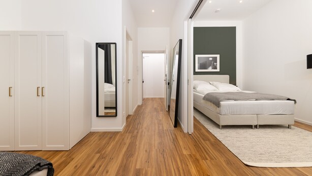 Een slaapkamer met een bed, een witte inbouwkast, een spiegel en een gang naar een andere kamer.