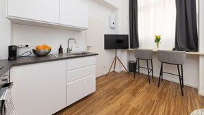 Een moderne keuken met witte kasten, een zwart aanrecht en een houten vloer. Er staat een schaal met sinaasappels, een koffiezetapparaat en een televisie op een statief. Twee barkrukken staan bij een raam met donkere gordijnen.