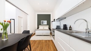 Een moderne, compacte hotelkamer met een houten vloer, een bed met witte lakens, een kitchenette en een eethoek met een donkere tafel en stoelen.