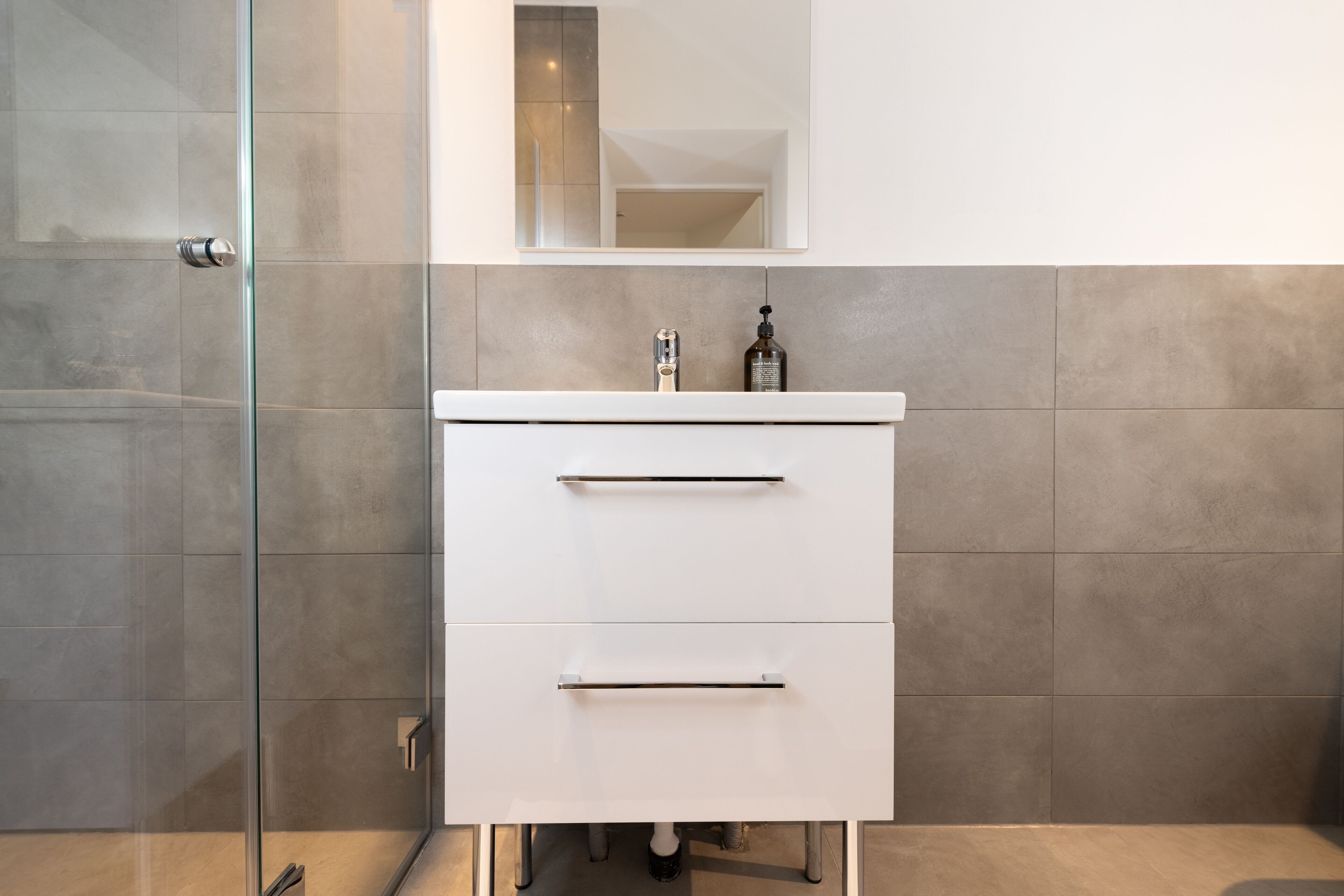 Une salle de bain moderne avec une douche à l’italienne en verre, un meuble-lavabo blanc doté de deux tiroirs et un miroir au-dessus du lavabo.