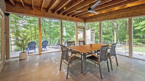 A covered outdoor dining area with a table and chairs, large windows, and a view of the surrounding forest. - Lakefront Roxboro Home w/ Private Dock! (Roxboro)