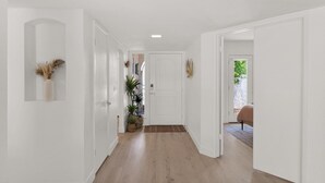 A hallway with white walls, wooden flooring, and a door leading to a bedroom with a bed and a view of greenery outside.
