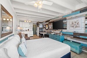A modern bedroom with a large bed, a desk, and a kitchenette in the background.