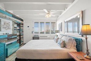 Un dormitorio con una cama, un ventilador de techo, una lámpara, una ventana con persianas y una pared con un diseño pintado.