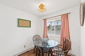 Een eetgedeelte met een glazen tafel, rieten stoelen, een plafondlamp, een raam met gordijnen en een lijst met een afbeelding aan de muur.