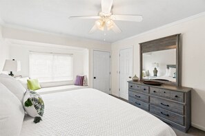 Een slaapkamer met een bed, een dressoir, een plafondventilator en een groot raam met jaloezieën.