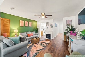 A living room with a green wall, a ceiling fan, a fireplace, and a kitchen in the background.