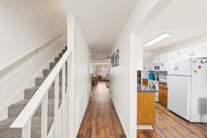 A hallway with a wooden floor, white walls, and a kitchen area with white cabinets and appliances.