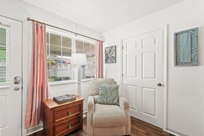 Una habitación con ventana, puerta, una silla, una mesita de noche y un adorno en la pared.