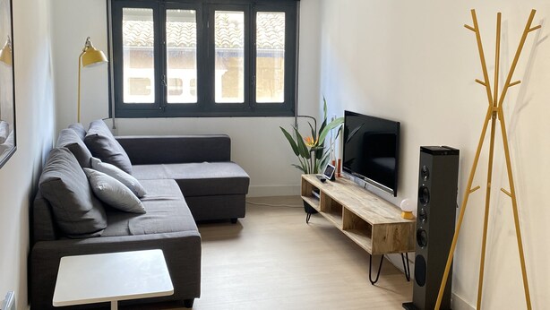 Un salon moderne avec un canapé d’angle gris, une petite table blanche, un téléviseur posé sur un meuble en bois et une grande fenêtre agrémentée de rideaux à motifs.