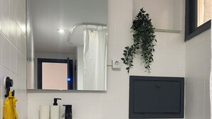 Une salle de bain avec un lavabo blanc, des toilettes et un miroir au-dessus du lavabo. Une serviette jaune est accrochée au mur, et une plante est posée sur le mur.