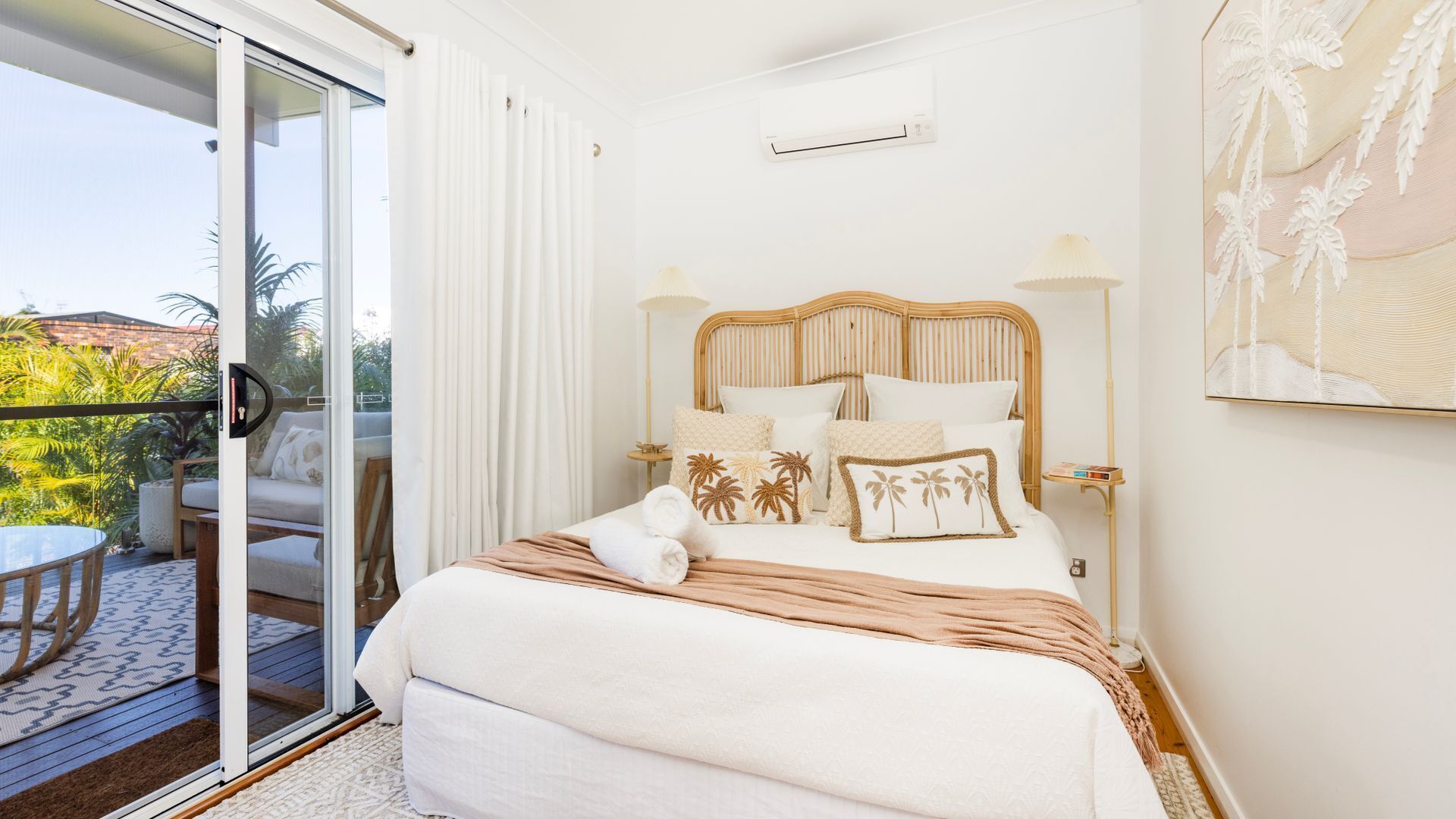 A bedroom with a bed, a bedside table, a lamp, a painting, and a sliding glass door to a balcony.