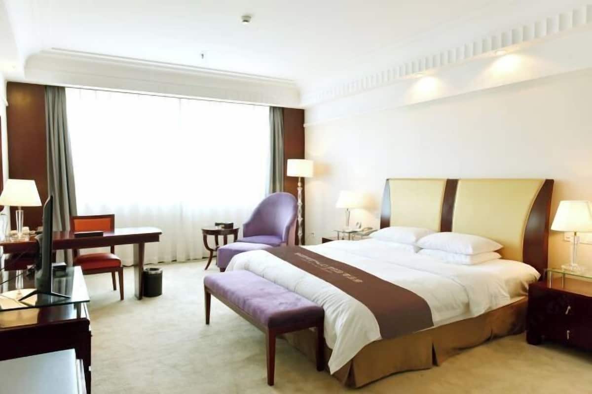 A hotel room with a large bed, a desk with a chair, a small table, and a bench.
