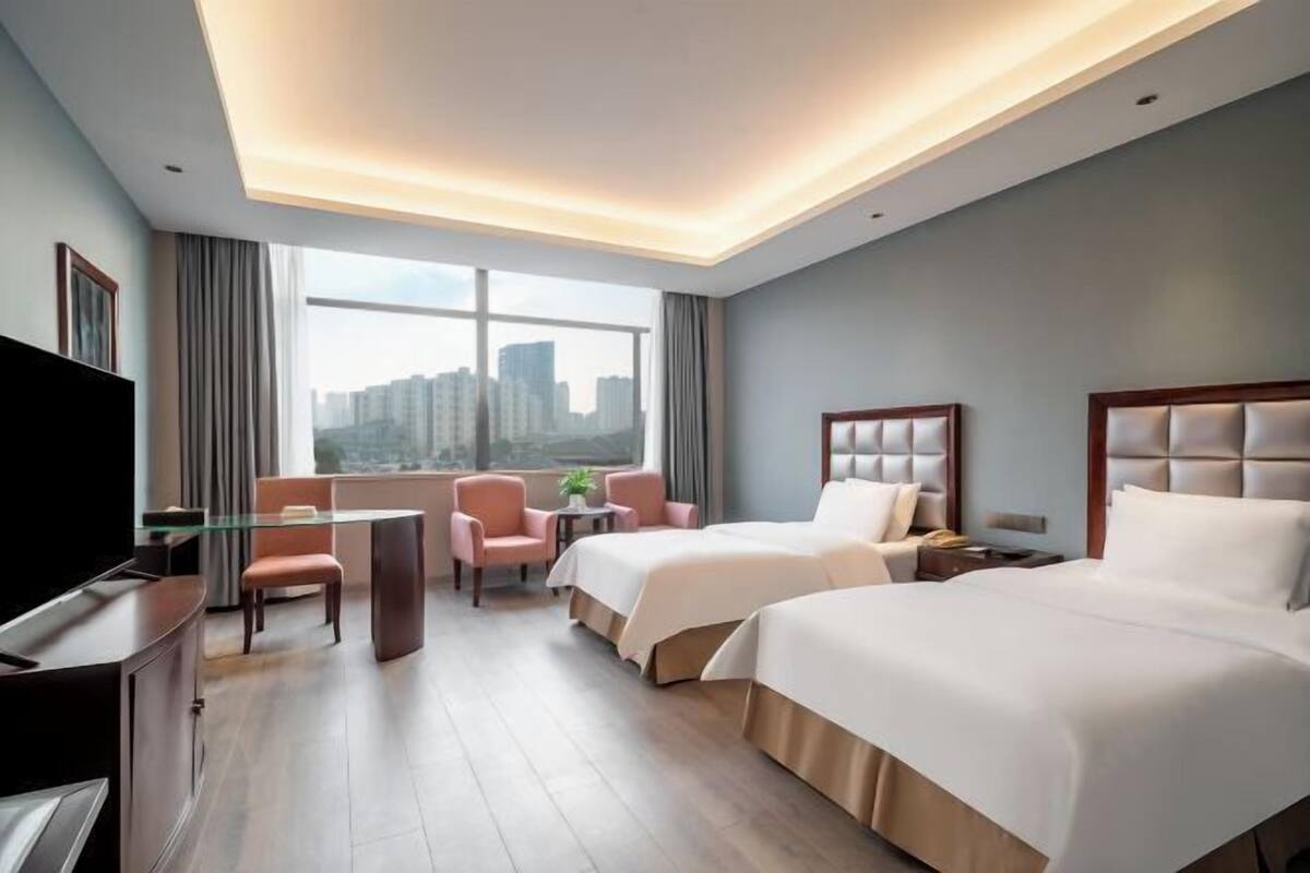 A hotel room with two beds, a desk, a TV, and a large window with a city view.